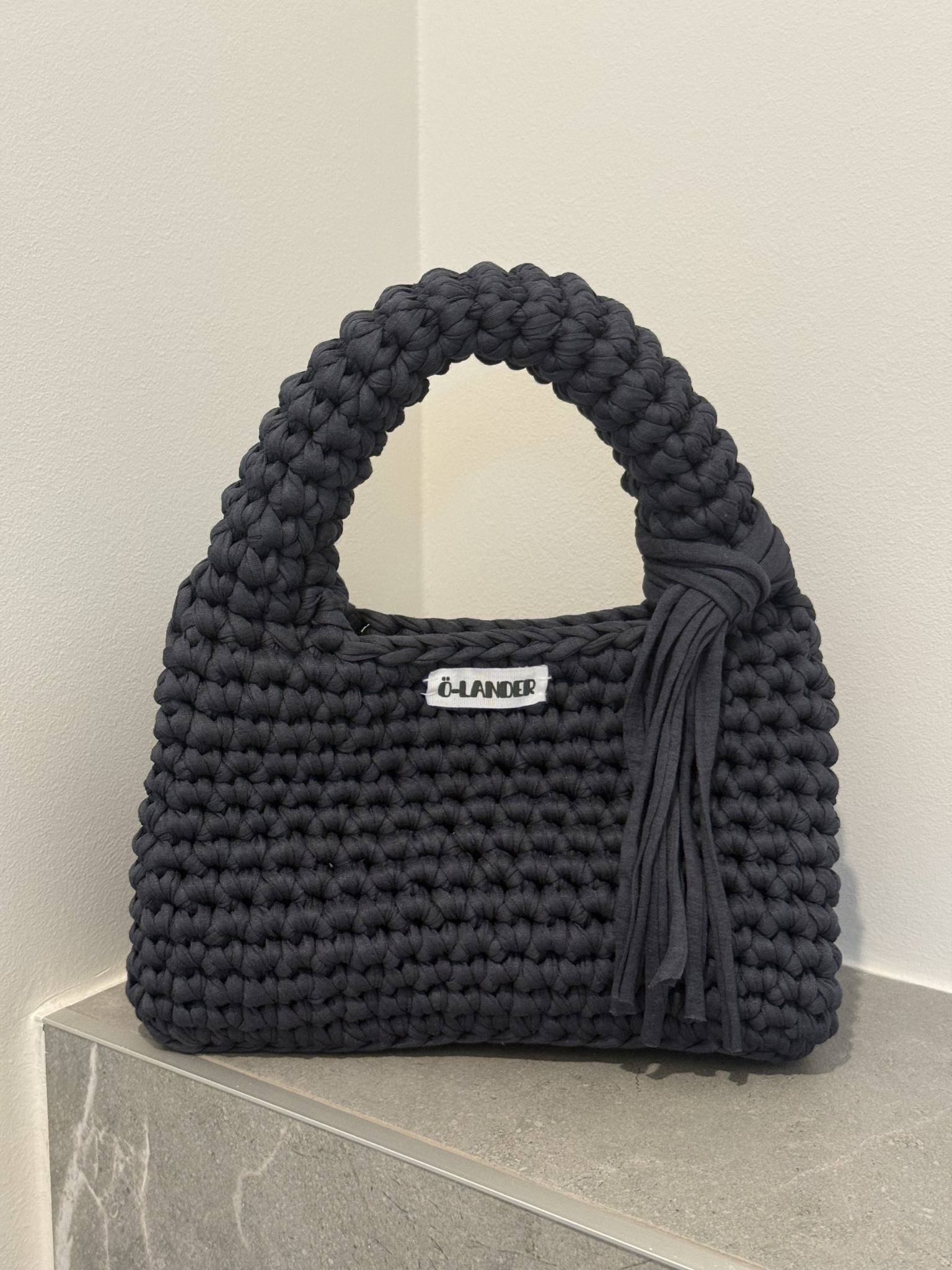 A stormy grey crocheted handbag with a top handle, a side tassel detail, and a small white brand label