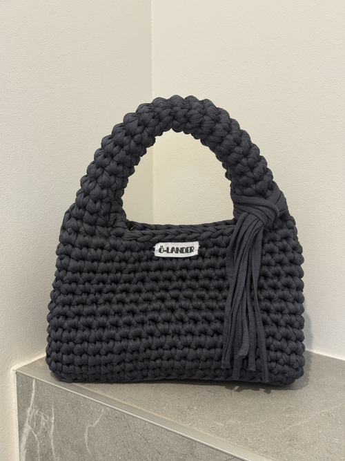 A stormy grey crocheted handbag with a top handle, a side tassel detail, and a small white brand label